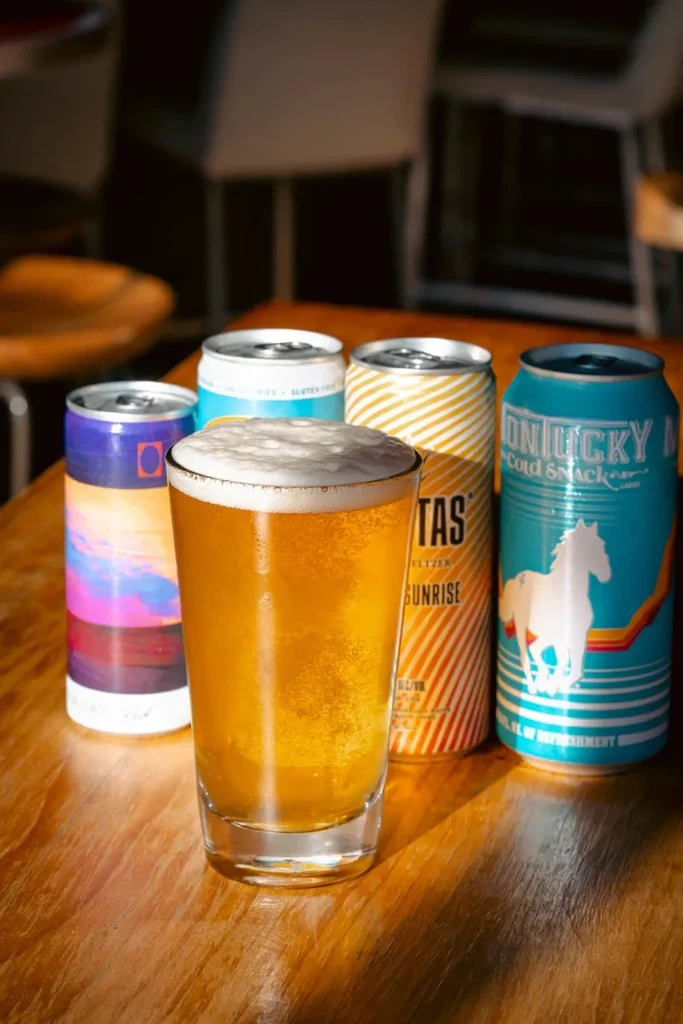 Amber beer with a foamy head, four brightly colored beverage cans.