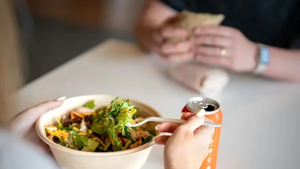 Hands hold a salad bowl and soda can at a table.