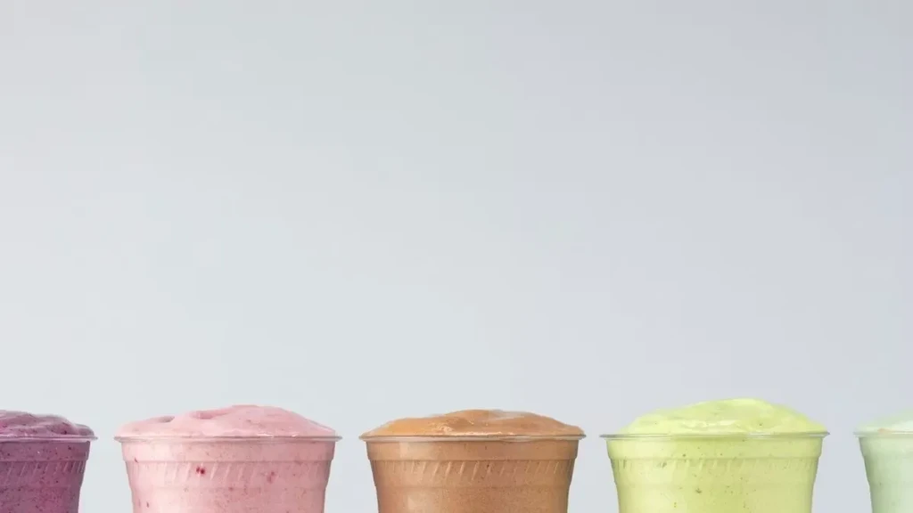 Five frothy, vibrantly colored smoothies in cups, plain grey background.