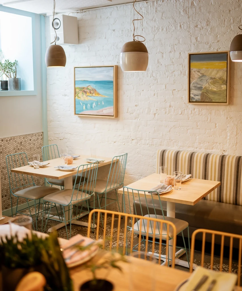 Restaurant interior with light wood tables, banquette, and pendant lights.