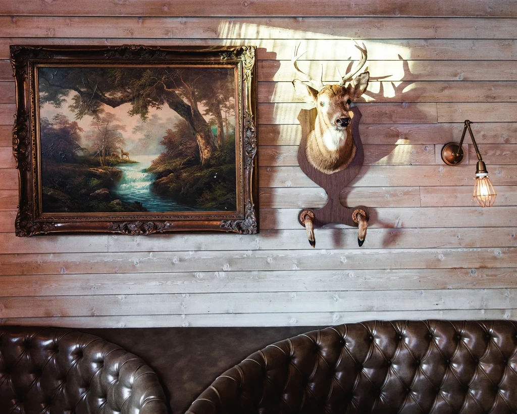 Rustic restaurant interior with framed forest painting and taxidermied deer head.