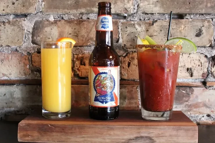 Three drinks on a wooden board: orange juice, beer, and Bloody Mary.