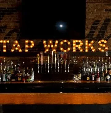Illuminated "TAP WORKS" sign above beer taps and liquor bottles.