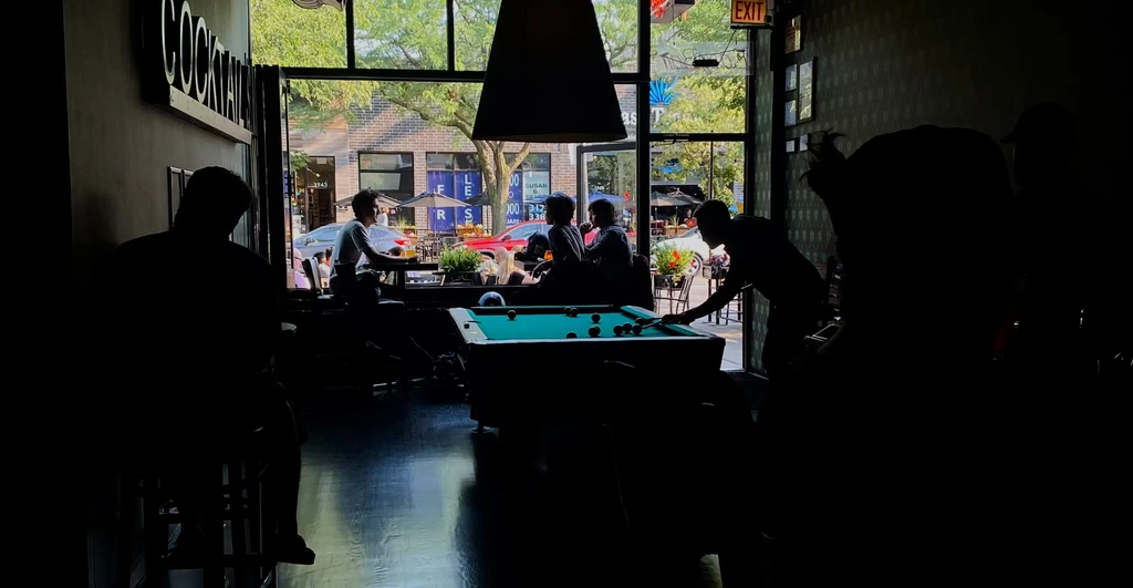 Dimly lit bar interior with silhouetted people and a pool table.