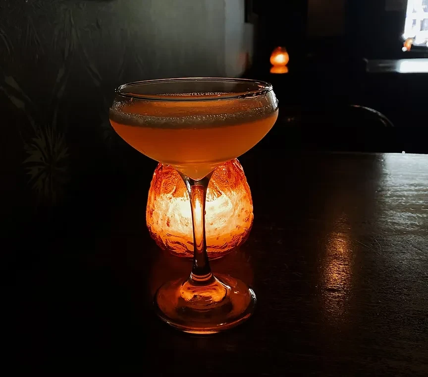 Orange-yellow cocktail in coupe glass, lit by a glowing candle.