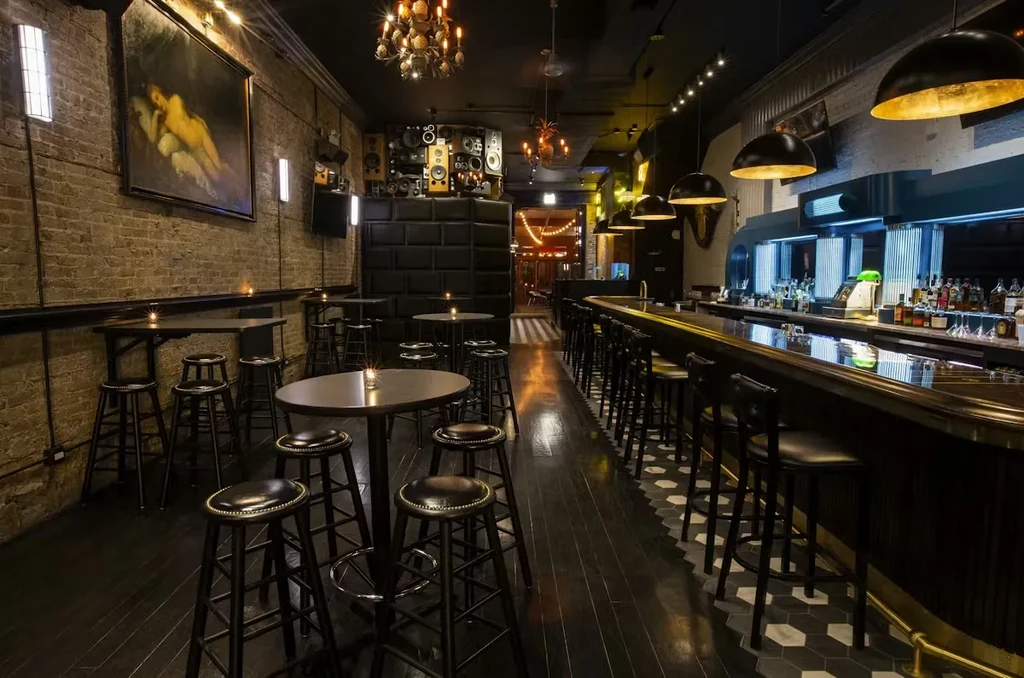 Dimly lit bar interior with stools, high-top tables, and unique chandeliers.