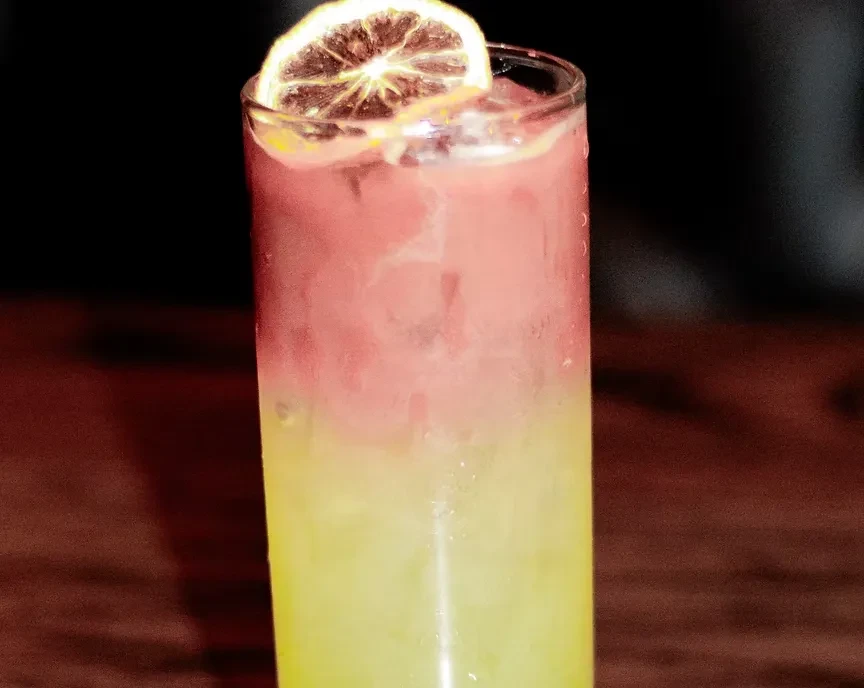 Layered pink and yellow drink with ice, garnished with citrus slice.