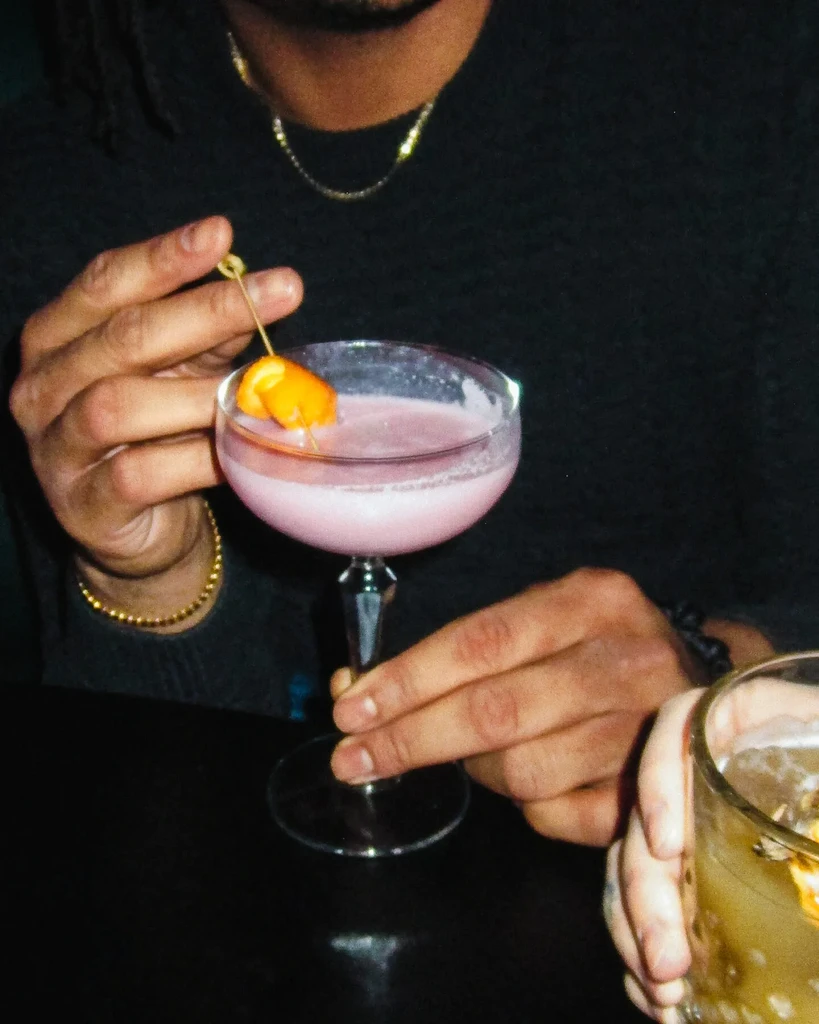 Hands hold a pink cocktail in a coupe glass.