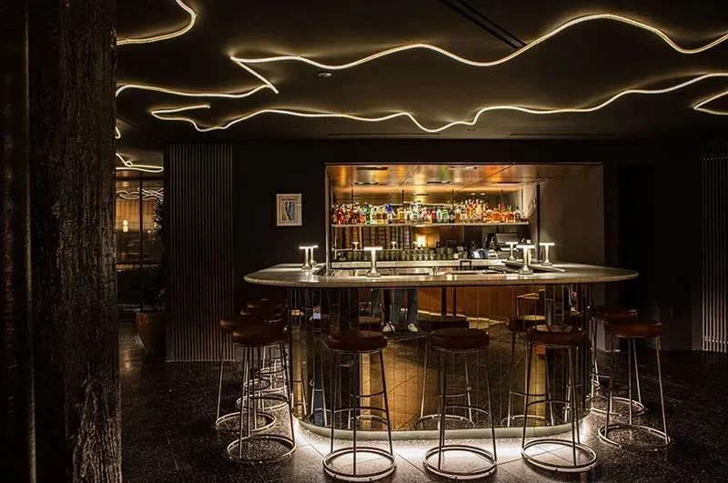 Modern bar interior with oval counter, stools, and backlit alcohol shelves.