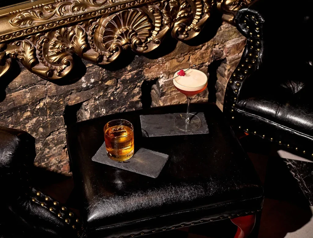 Two cocktails with ice and garnish on coasters and a dark table.