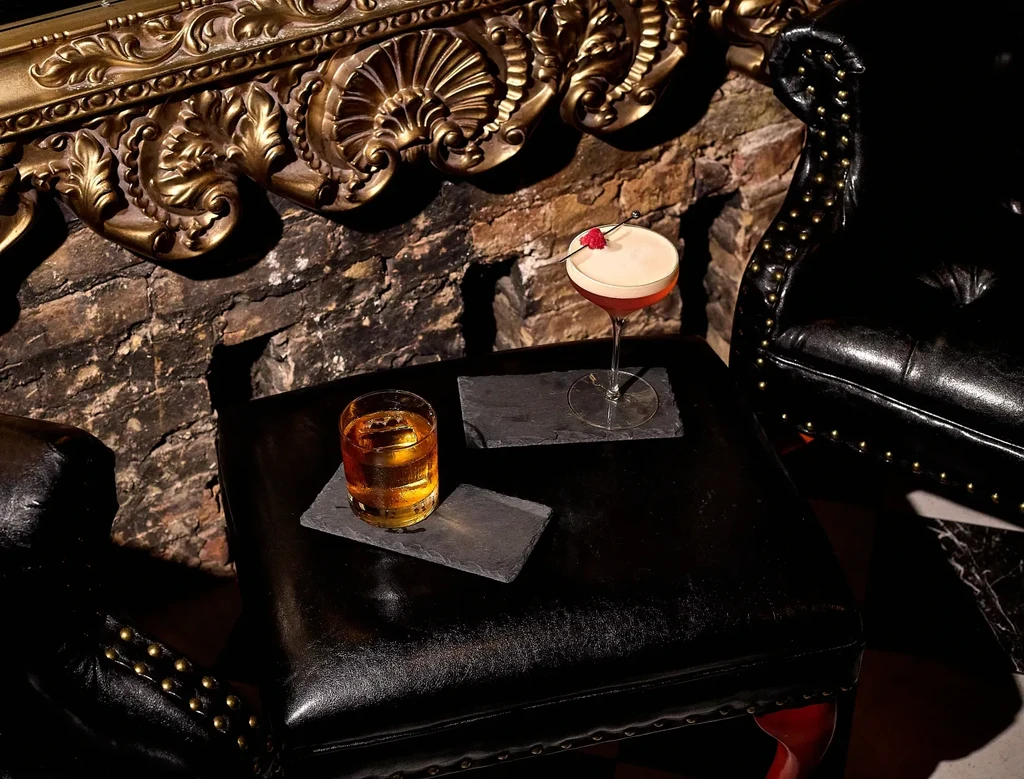 Two drinks on coasters sit on a dark ottoman by a brick wall.