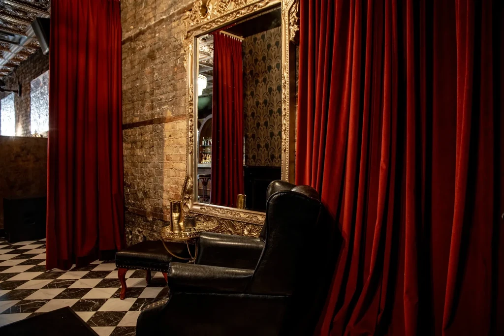 Ornate lounge with red velvet curtains, mirror, leather armchair, checkered floor.