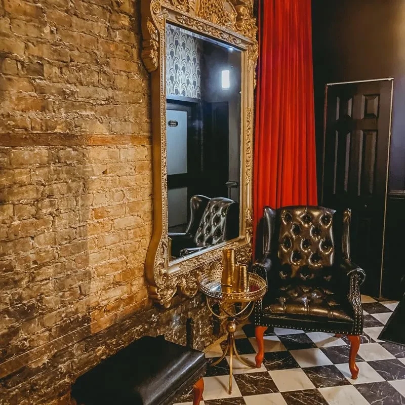 Ornate gold mirror, leather armchair, and checkered floor in a dimly lit room.