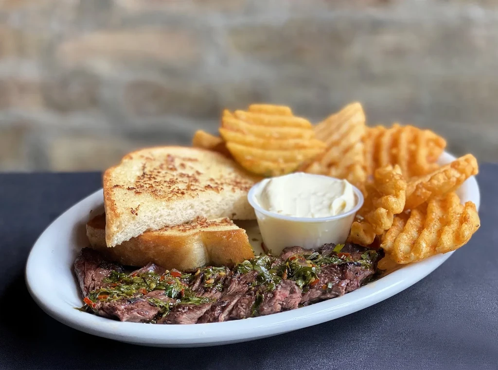 Sliced steak, garlic toast, waffle fries, and dipping sauce on a plate.