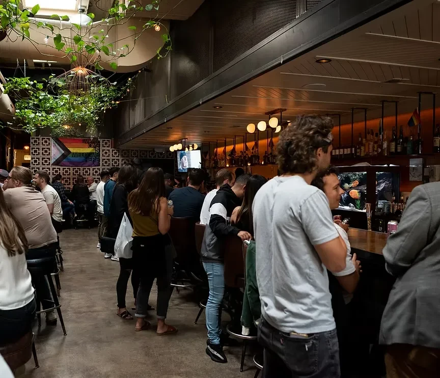 Crowded bar interior with patrons, plants, and a "Pride" flag sign.