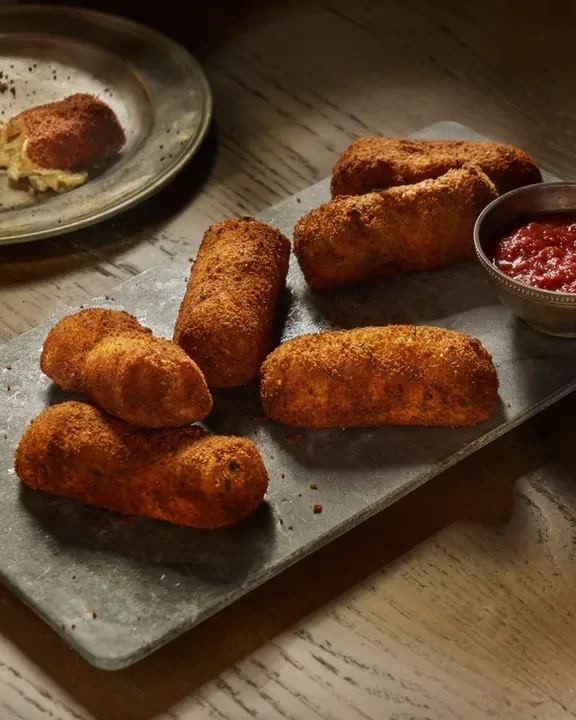 Six croquettes on a slate, with marinara sauce and crumbs.
