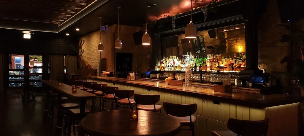 Dimly lit bar interior with a long wooden counter and many bottles.