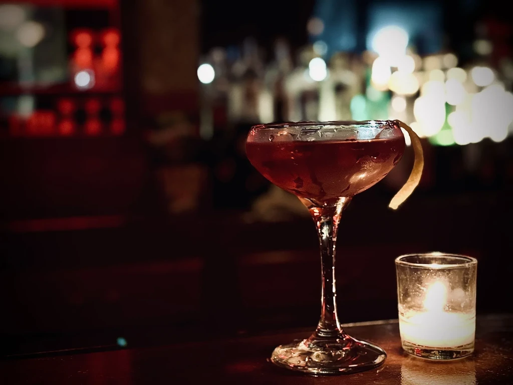 Red cocktail in coupe glass with lemon peel and votive candle.