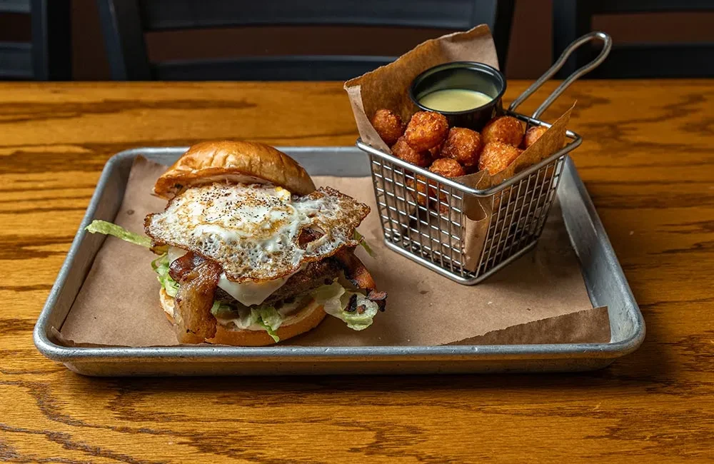 Gourmet burger, tater tots, and dipping sauce on a metal tray.