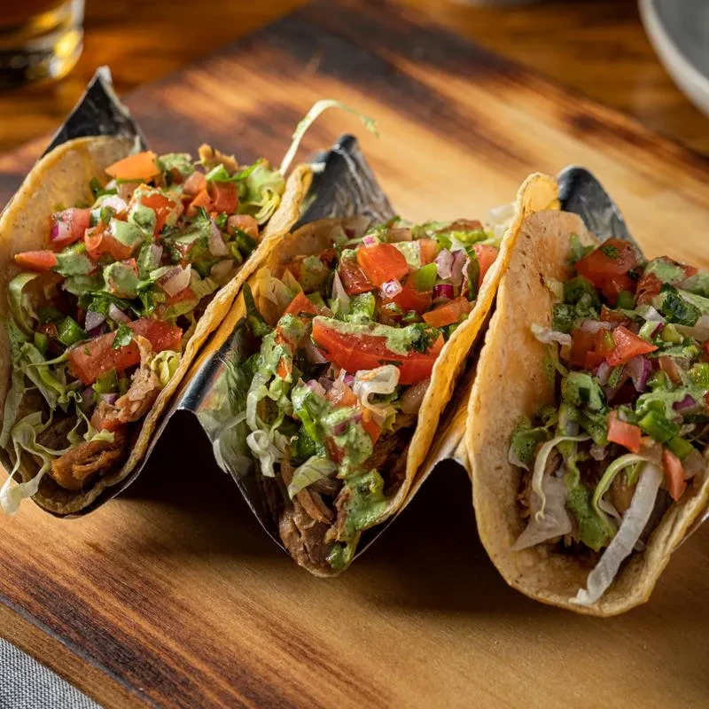 Three vibrant tacos in a metal holder on a wooden board.