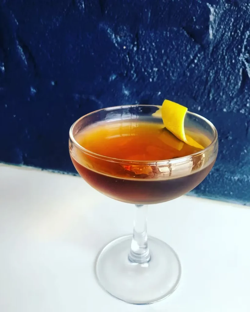Amber cocktail in coupe glass with lemon peel on white surface.