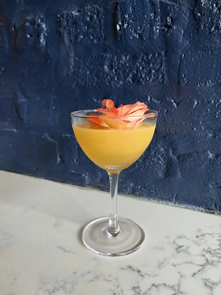 Cocktail in a coupette glass garnished with a peach-colored flower.