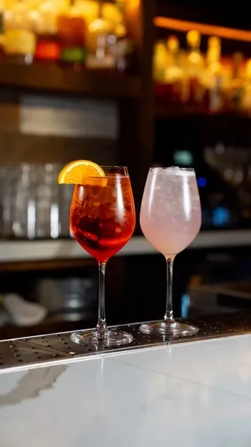 Two stemmed drinks with ice and garnishes on a bar.