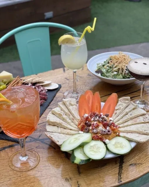 Rustic table with three cocktails, hummus plate, and charcuterie.