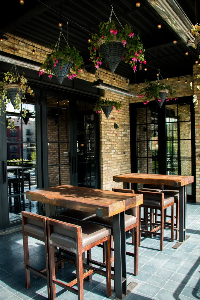 Outdoor patio with high-top tables, stools, and hanging plants.