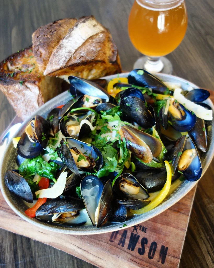 Steamed mussels in broth with bread and beer on a wooden surface.
