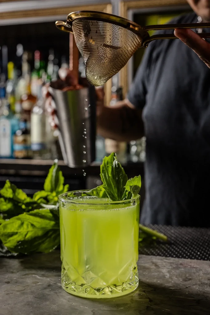 Vibrant green cocktail being strained into a textured glass.