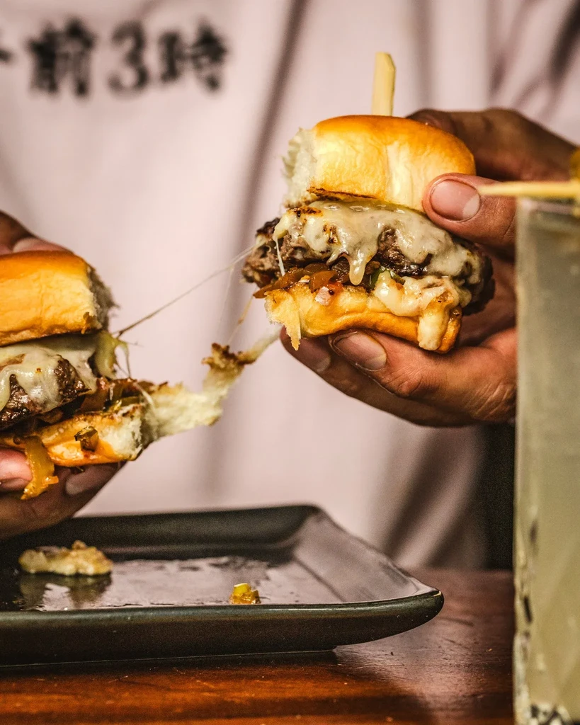 Hands holding two mini-burgers, one with a string of melted cheese.