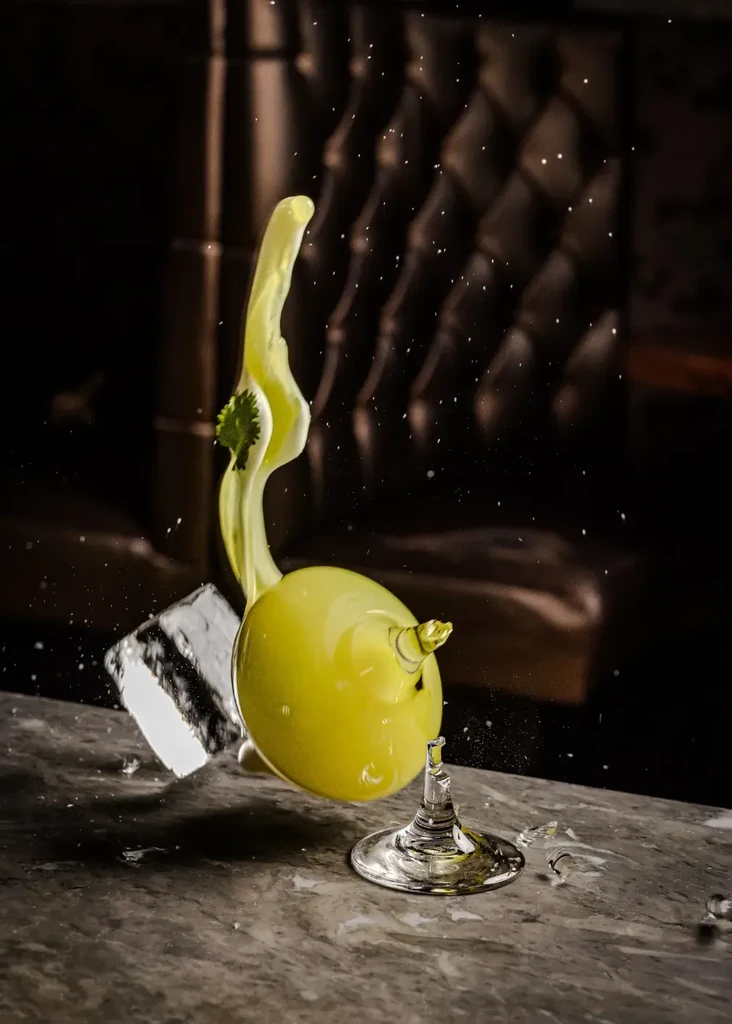 Green cocktail spills from a broken glass onto a dark surface.