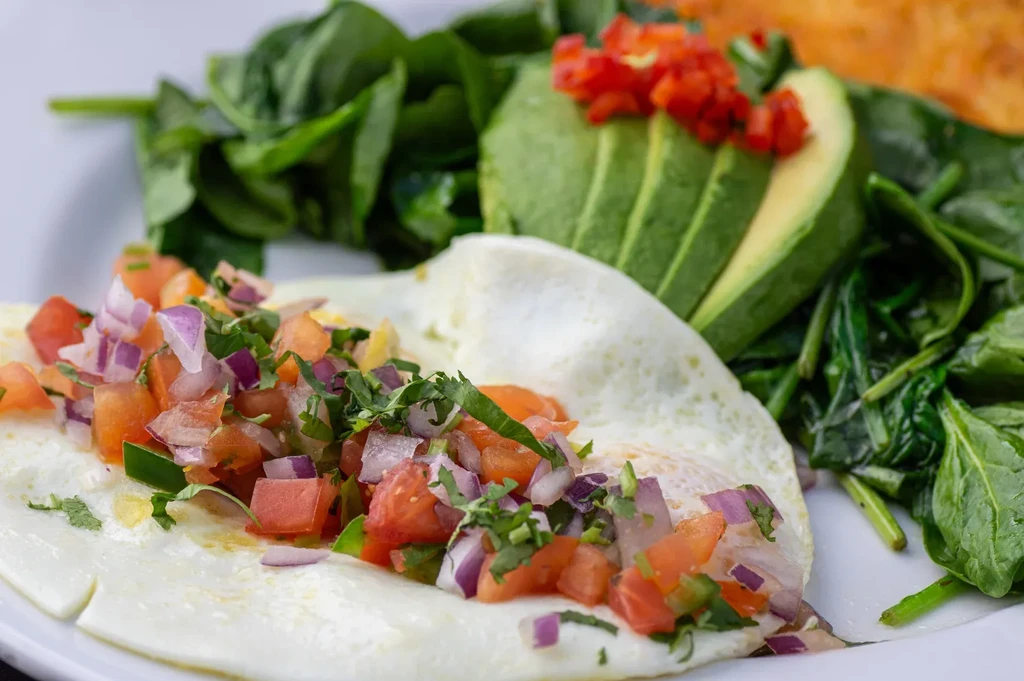 Fried eggs with pico de gallo, avocado, and spinach on a white plate.