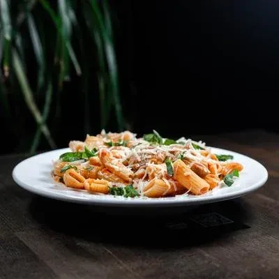 Penne pasta with reddish sauce, cheese, and herbs on a dark table.