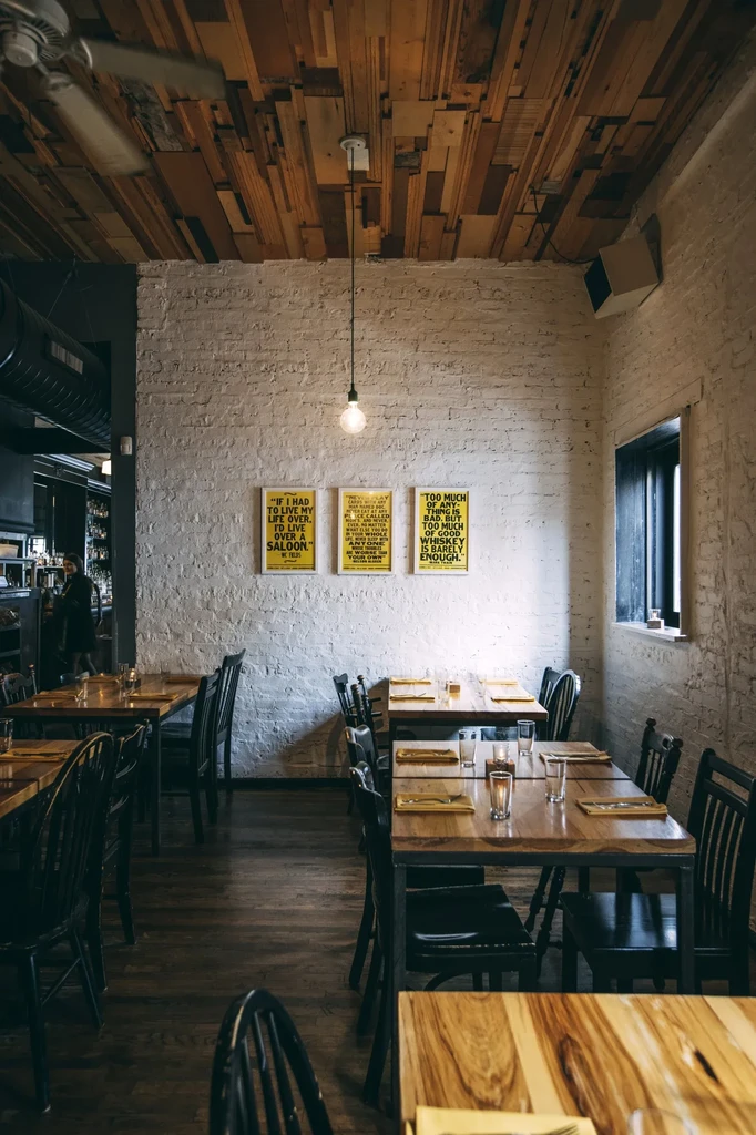 Restaurant interior with wooden tables, brick wall, and mosaic wood ceiling.