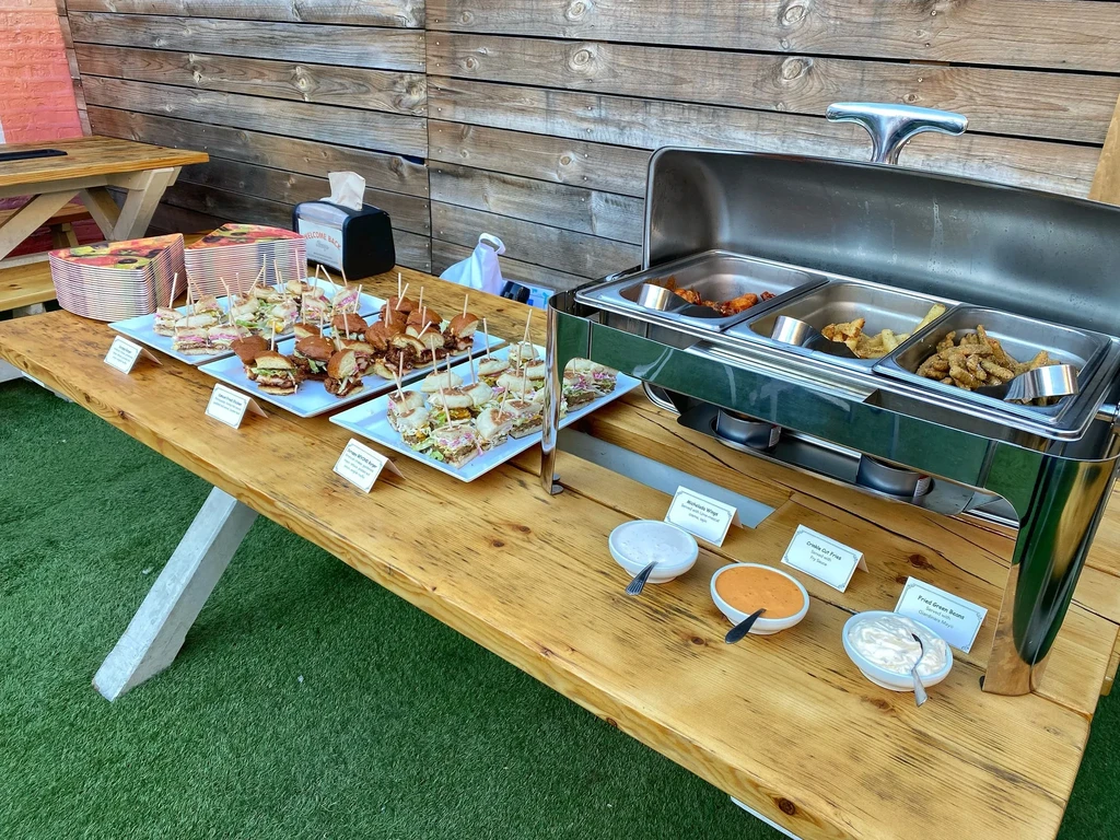 Buffet table with mini sandwiches, hot food, and sauces outdoors.