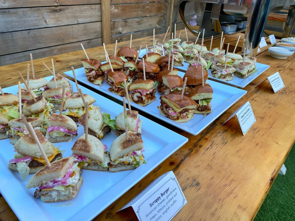 Buffet of sliders with cheese, lettuce, pickled onions, and skewers.