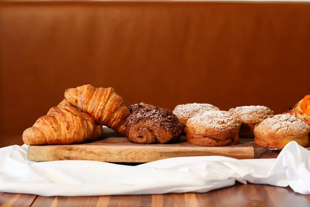 Pastries on a wooden cutting board, including croissants, muffins.