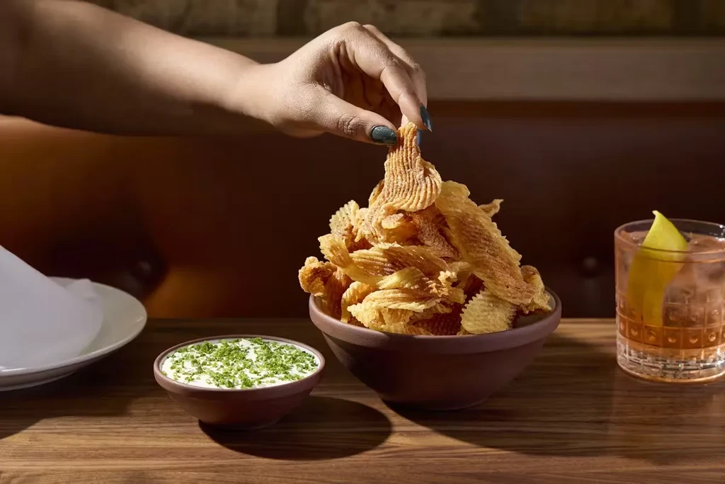Hand dips into a bowl of chips next to a cocktail.