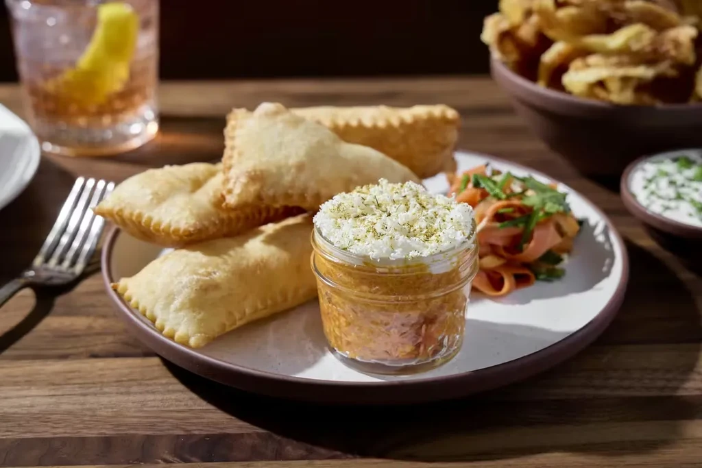 Fried pastries with dip, sliced vegetables, and pink drink on table.