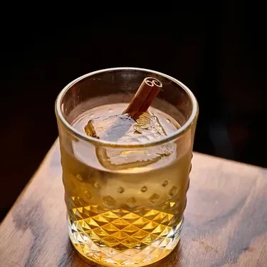 Amber drink in textured glass with ice cube and cinnamon stick.