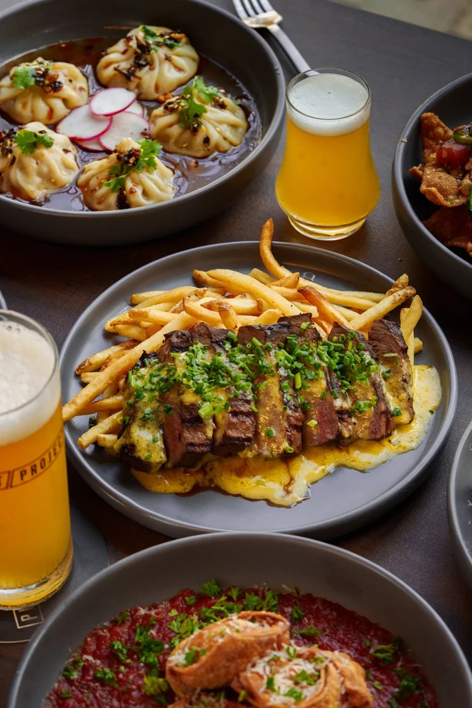Grilled steak, fries, dumplings, and beer on a dark table.
