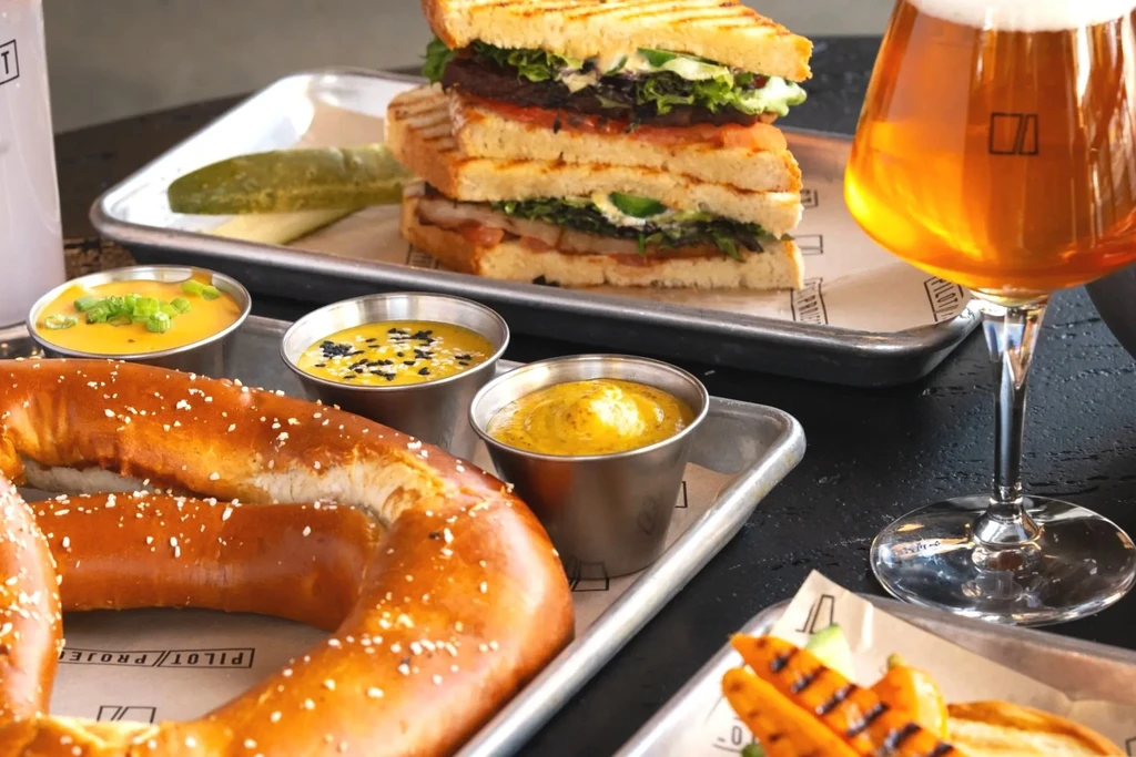 Pretzel, sauces, stacked sandwich, pickle, and beer on a table.