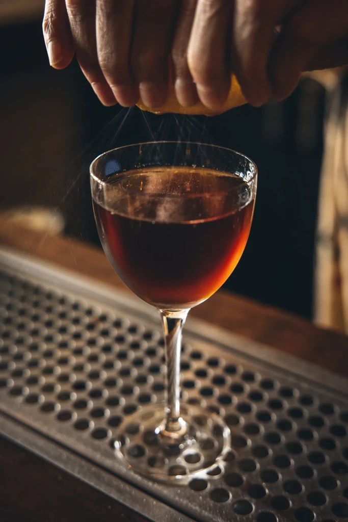 Hands squeezing citrus peel over a dark cocktail in a stemmed glass.