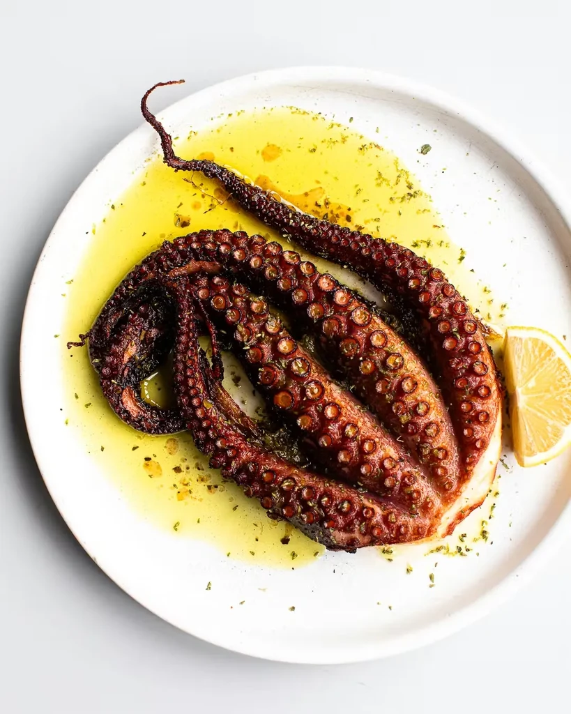 Cooked octopus dish glistening with oil, herbs, and a lemon wedge.