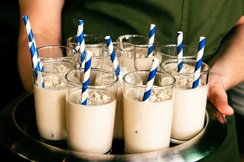 Eight creamy drinks with striped straws on a metallic tray.