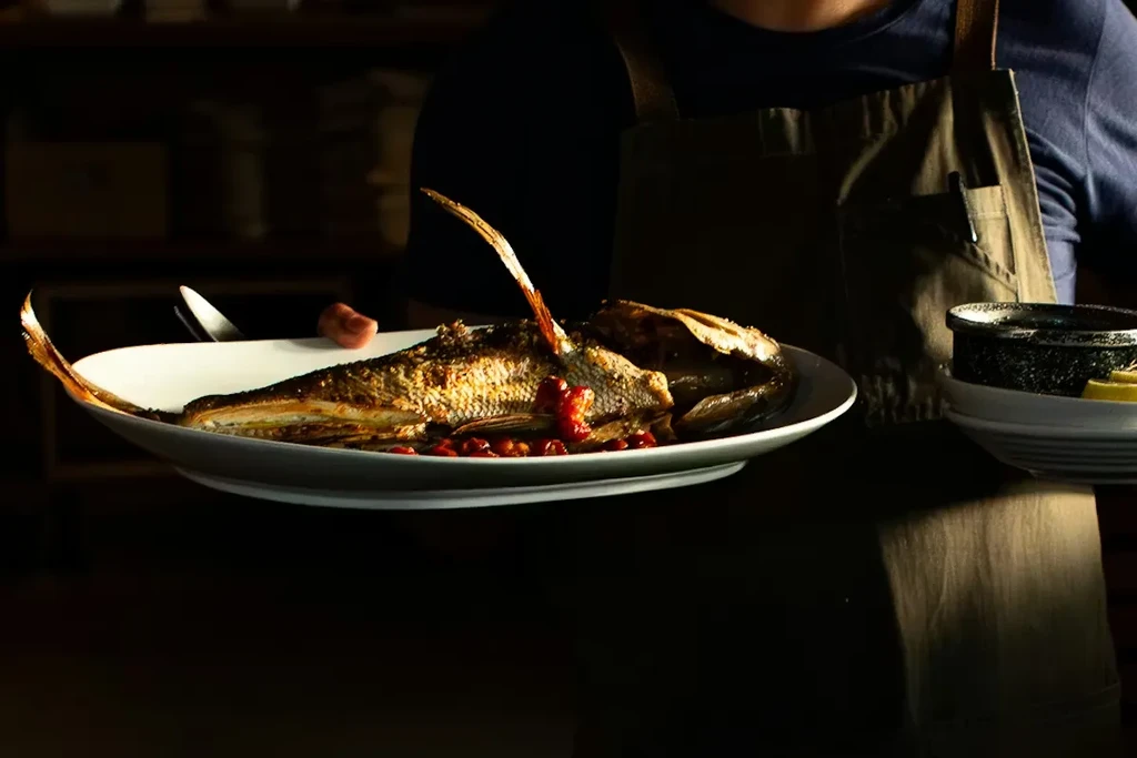 Whole grilled fish on a white plate with lemon wedges.
