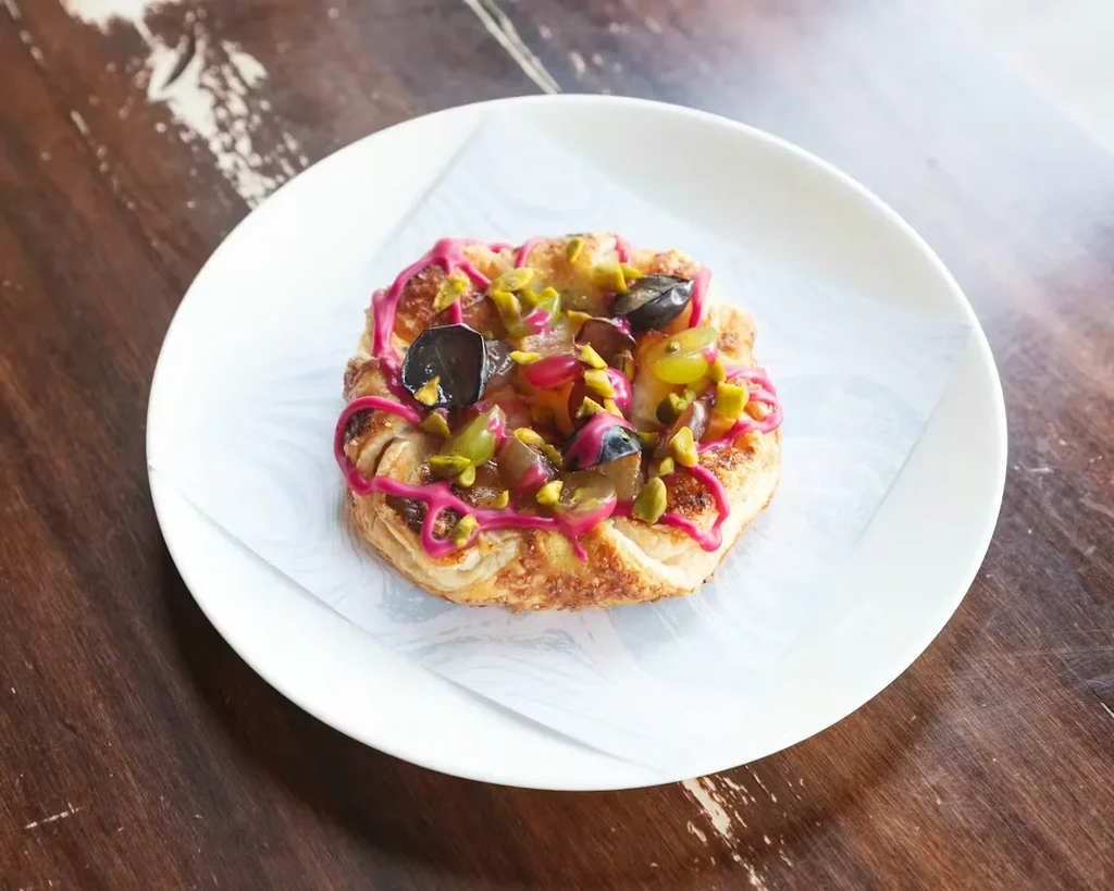Round pastry with fruit, pistachios, and pink icing on a plate.