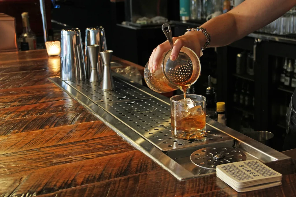 Bartender pouring a light brown cocktail into an ice-filled glass.